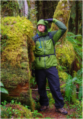 Picture of Brian standing next to a fallen tree, the diameter of which is the same as Brian’s height. Everything is covered in bright green moss, the color of Brian's raincoat as well.