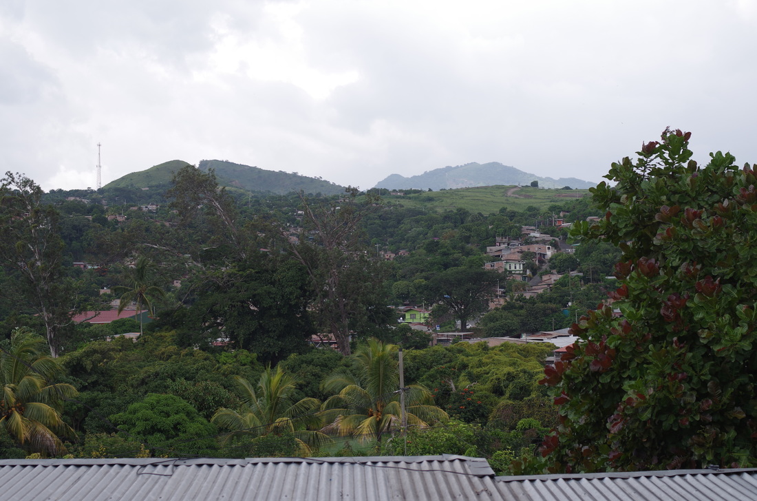 Picture: a landscape, tin roofs in sight at the bottom, trees and houses scattered throughout the valley, mountains er more like large hills rise on the other side of the valley giving way to a gray sky signalling impending rain
